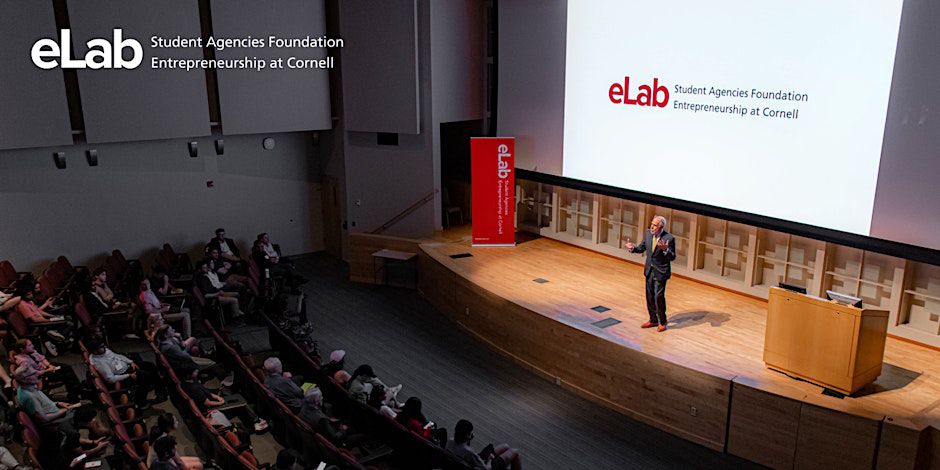 An old Caucasian man in suits speaking on stage in an auditorium filled with people.