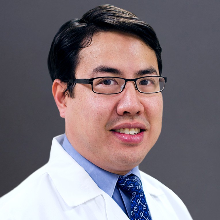 Young East Asian man wearing glasses, a dark blue tie with white design, and a white coat