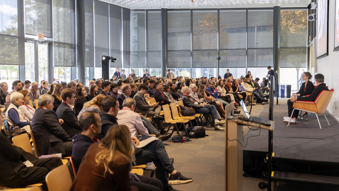 A Caucasian couple sitting on a stage in a room filled with audience