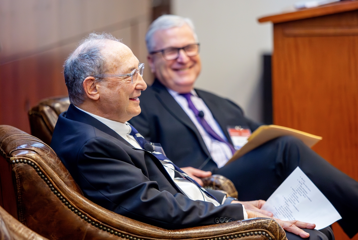 Two older men wearing suits and ties smiling and talking.