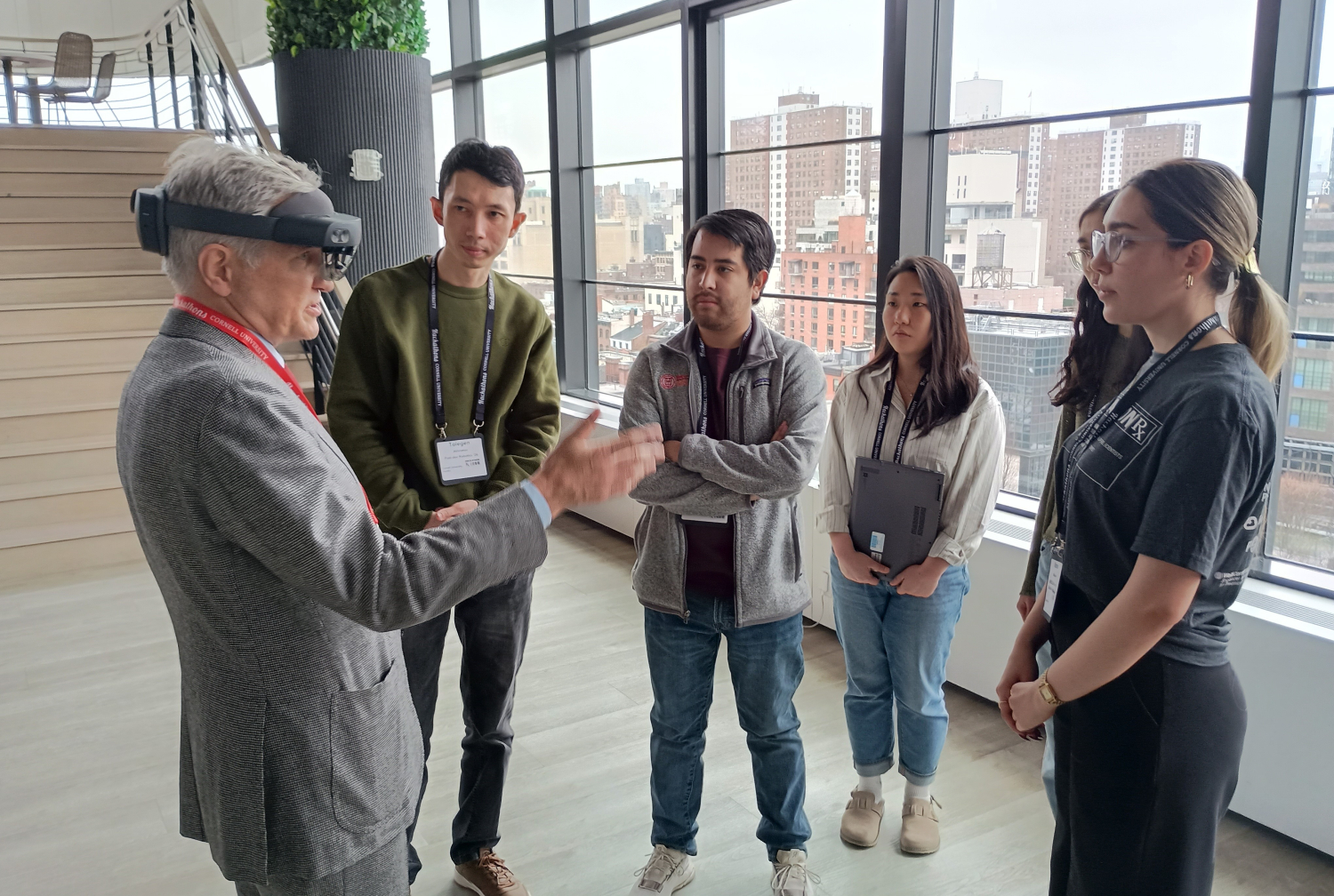An old man wearing VR goggles and a gray suit talking to a group of students standing in a semicircle 