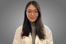 An East Asian woman with long hair and glasses wearing a gray sweater and white coat