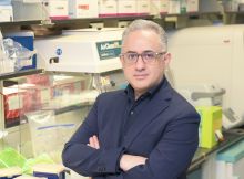 A male scholar with glasses and grey hair. He's wearing a black shirt and dark blue suit posing in a biology lab.