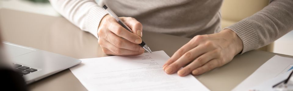 Two hands on a table.  A person is filling out a form with a pen in their right hand. 