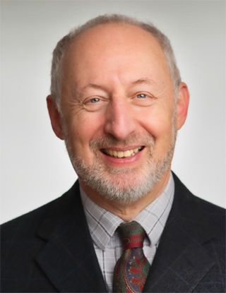An older Caucasian man with a beard wearing a black suit and a gray tie with red flowers